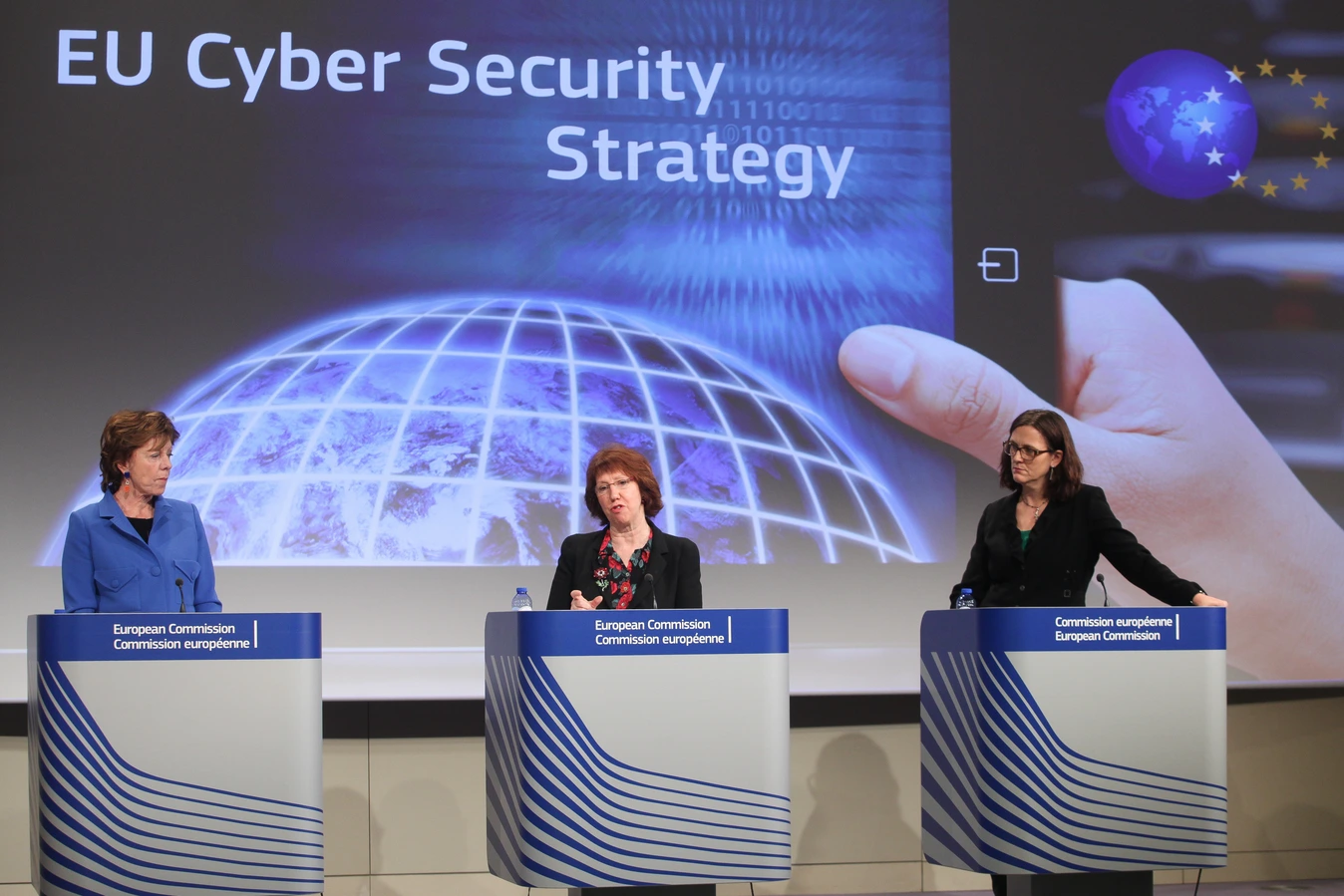 Pressekonferenz zur EU Cyber-Sicherheitsstrategie, mit drei Rednern an Podien vor einem Bildschirm, der die Strategie zeigt.